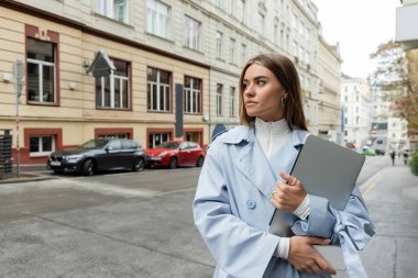 pretty woman in blue trench coat holding smartphone and laptop while walking on street in Vienna  clipart