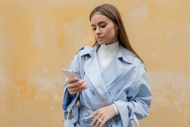 Mavi trençkotlu genç kadın Viyana caddesinde yıpranmış sarı duvarın yanında akıllı telefon kullanıyor. 