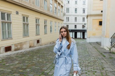 young woman in blue trench coat talking on smartphone while walking on street in Vienna  clipart