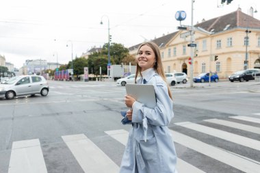 Mavi trençkotlu mutlu kadın elinde dizüstü bilgisayarla Viyana 'da karşıdan karşıya geçiyor. 
