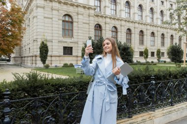 Mavi trençkotlu mutlu kadın elinde dizüstü bilgisayarla Viyana 'daki tarihi binanın yakınındaki akıllı telefondan fotoğraf çekiyor. 