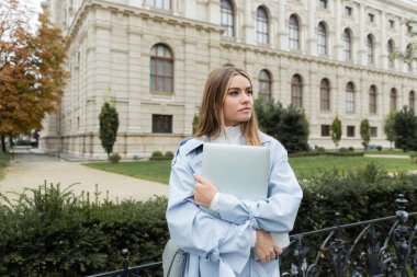 Viyana 'daki tarihi binanın yanında elinde dizüstü bilgisayar tutan mavi trençkotlu güzel genç bir kadın. 