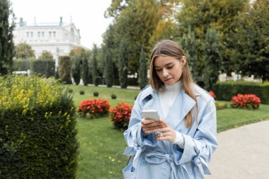 Viyana 'da yeşil parkta dikilirken akıllı telefon kullanan mavi trençkotlu genç kadın. 