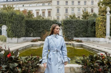 young woman in blue trench coat standing near fountain and historical building in green park in Vienna  clipart