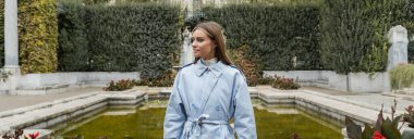 young woman in blue trench coat standing near fountain with green water in park, banner  clipart