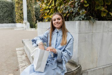 smiling young woman in blue trench coat sitting with shopping bag on concrete bench in green park in Vienna  clipart