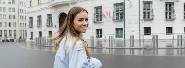 positive young woman in blue trench coat smiling on urban street in Vienna, banner  clipart