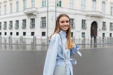 pleased young woman in trendy and blue trench coat smiling on urban street in Vienna  clipart