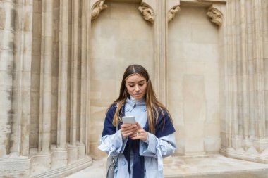 Mavi trençkotun üstünde fuları olan genç bir kadın Viyana 'daki tarihi binanın yanındaki akıllı telefonu kullanıyor. 