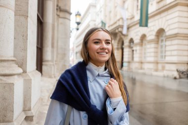 happy and stylish woman with scarf on top of blue trench coat smiling while looking away on street in Vienna clipart