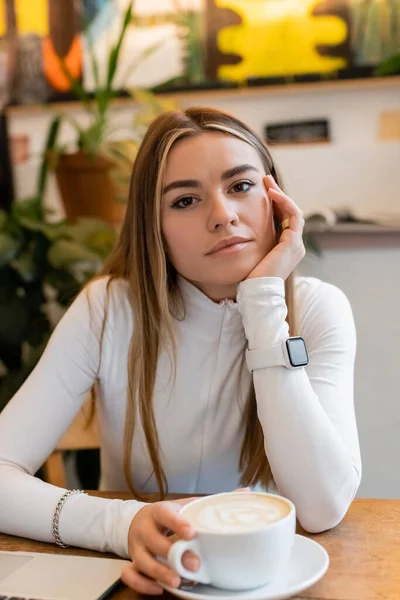 young woman in turtleneck with smart watch sitting next to cup of cappuccino in cafe