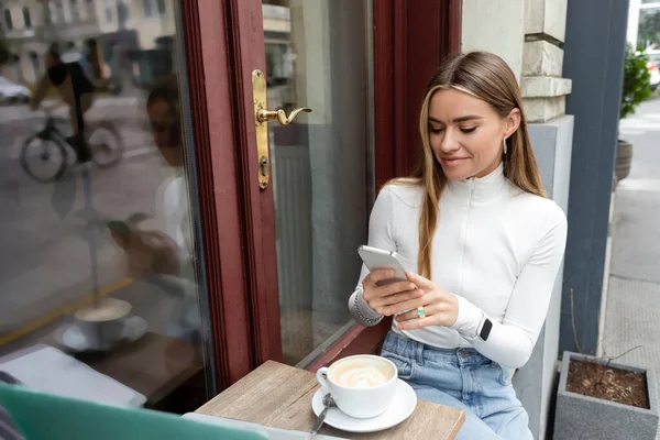 ウィーンのカフェに座っている間にカプチーノのカップの近くでスマートフォンを使って笑顔の若い女性 — ストック写真