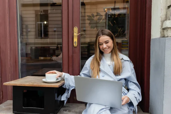 happy woman in blue trench coat using laptop while reaching cup of coffee in outdoor cafe in Vienna