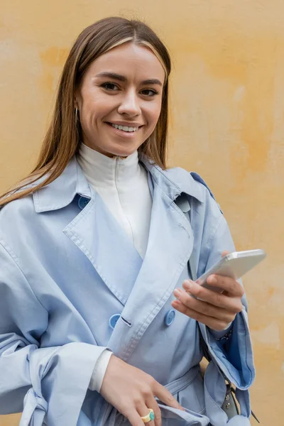 在维也纳的大街上 身穿蓝色战壕外套的快乐女人拿着智能手机站在黄色的墙边 — 图库照片
