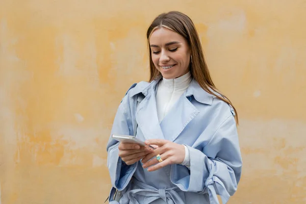 幸せな女性で青トレンチコートメッセージングでスマートフォン近く風化黄色の壁に路上でウィーン — ストック写真