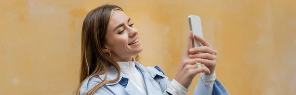 Lycklig Kvinna Blå Trenchcoat Foto Smartphone Nära Väderbiten Gul Vägg — Stockfoto