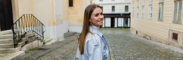 青いトレンチコートを着た陽気な若い女性がウィーンの通りの建物の近くを歩いていて — ストック写真