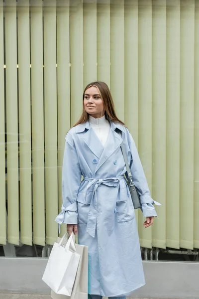青いトレンチコートを着た可愛い女性が紙袋を持ってウィーンの通りの窓の近くに立っている間 — ストック写真