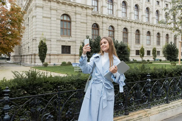 在维也纳历史建筑附近 身穿蓝色风衣的快乐女人拿着笔记本电脑 在智能手机上拍照 — 图库照片