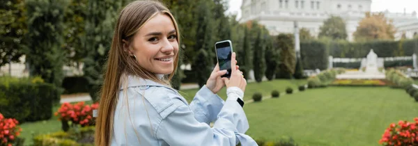 快乐的年轻女人穿着蓝色的风衣 在智能手机和横幅上为绿色公园拍照 — 图库照片