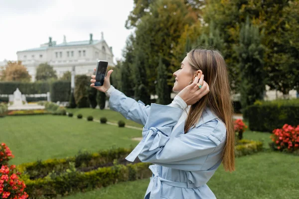 携帯電話で自撮りしながらウィーンの緑の公園に立つブルートレンチコートの陽気な女性 — ストック写真