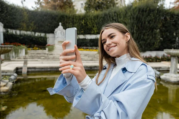 身穿蓝色风衣的年轻女子微笑着在公园喷泉边的智能手机上拍照 — 图库照片