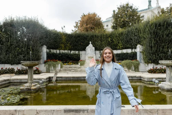 緑の公園の噴水の近くで手を振って青いトレンチコートの若い女性の笑顔 — ストック写真
