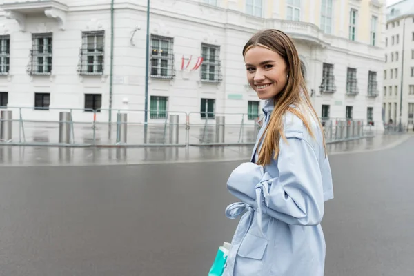 Mavi trençkotlu tatmin olmuş kadın Viyana 'da şehir caddesinde gülümsüyor. 
