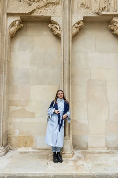ウィーンの歴史的建造物の近くに立つ青いトレンチコートの上にスカーフを持つ若い女性 — ストック写真