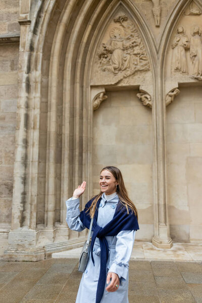 веселая молодая женщина с шарфом на верхней части голубой траншеи машет рукой возле исторического здания в Вене 