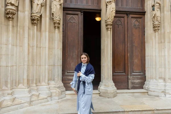 青いトレンチコートの上にスカーフを持つ若い女性は ウィーンの歴史的建造物の近くを歩く — ストック写真
