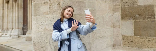 青いトレンチコートの上にスカーフを持つ幸せな女性は ウィーンの歴史的な建物の近くにビデオ通話を持っています バナー — ストック写真