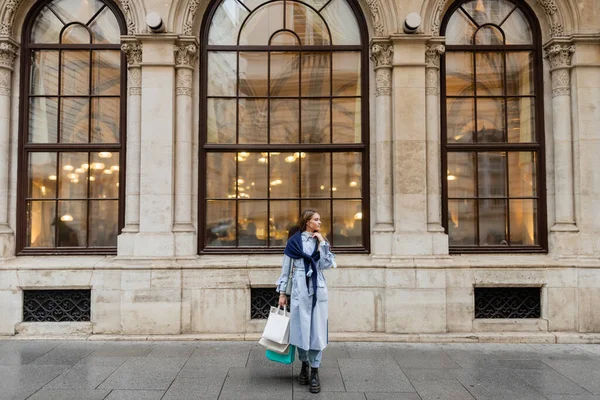 young and stylish woman with scarf on top of blue trench coat holding shopping bags near historical building in Vienna