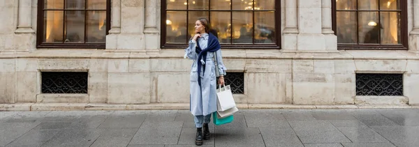 young and stylish woman with scarf on top of blue trench coat holding shopping bags near historical building in Vienna, banner