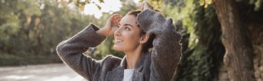 Smiling brunette woman in warm jacket touching hair and looking away while spending time and relaxing in blurred park at daytime in Barcelona, Spain, banner  clipart