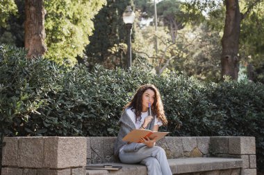Pensive young and curly woman in casual clothes holding notebook and marker near lips while sitting near devices on stone bench and green plants in park at daytime in Barcelona, Spain  clipart