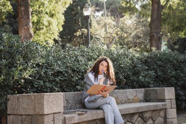 Focused young curly woman holding marker pen near lips and notebook while looking at camera near gadgets on stone bench and green plants in park in Barcelona, Spain  clipart