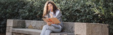 Focused young curly woman looking at camera while holding marker and notebook near smartphone and laptop on stone bench near bushes in park in Barcelona, Spain, banner, work from anywhere  clipart