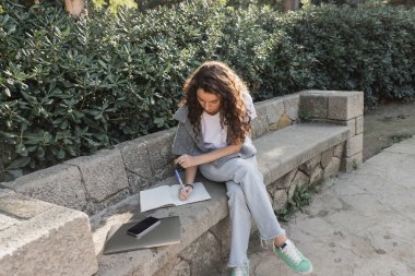 Young and curly woman in warm sweater on shoulders writing on notebook near laptop and smartphone with blank screen on stone bench beside green bushes in park in Barcelona, Spain  clipart