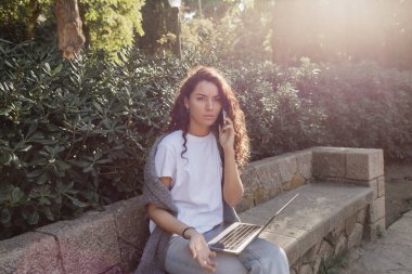 Pensive young and curly freelancer in casual clothes talking on smartphone near laptop while sitting on stone bench and spending time near green plants in park in Barcelona, Spain  clipart