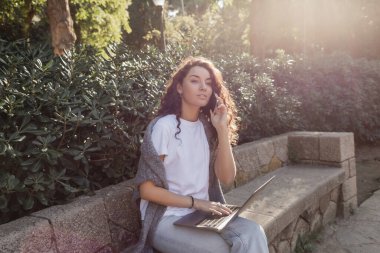 Young and curly freelancer in casual clothes talking on smartphone and looking away while using laptop and sitting on stone bench in green park in Barcelona, Spain  clipart