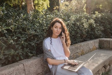 Barcelona, İspanya 'da park halindeki yeşil çalılıkların yanındaki taş bankta kameraya bakarken akıllı telefondan konuşan ve laptop kullanan genç ve kıvırcık serbest çalışan. 