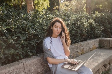 Young and curly freelancer in casual clothes talking on smartphone and using laptop while looking at camera on stone bench near green bushes in park in Barcelona, Spain  clipart