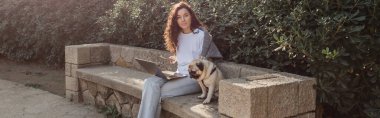Genç ve kıvırcık güzel bir kadın kameraya bakıp Barselona, İspanya 'daki yeşil parktaki taş bankta vakit geçirirken aletler kullanıyor ve köpeğini okşuyor. 