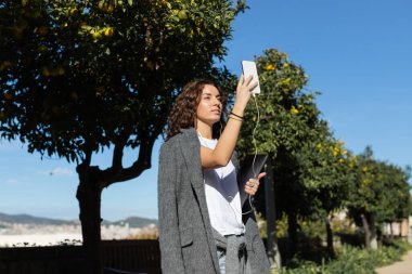 Young and curly freelancer in warm jacket and casual clothes using earphones and smartphone and holding laptop while standing near trees in park in Barcelona, Spain  clipart