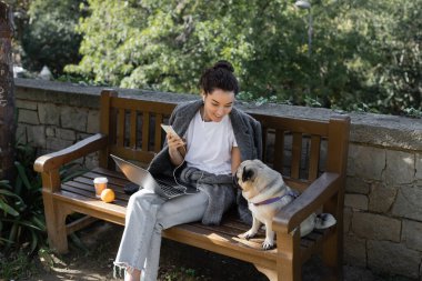 Kulaklıkla gülümseyen, akıllı telefon tutan ve dizüstü bilgisayarın yanında oturan köpek yavrusuna bakan, gitmek için kahve içen, ahşap bankta portakal yiyen ve İspanya 'nın Barcelona kentinde parkta vakit geçiren biri. 