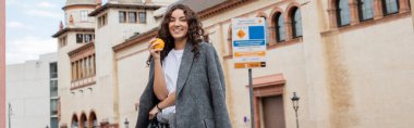 Gri ceketli genç kıvırcık kadın olgun ve taze portakallı ve kameraya gülümseyen Barselona, İspanya 'nın arka planında tarihi bir dönüm noktası olan afiş, antik bina 