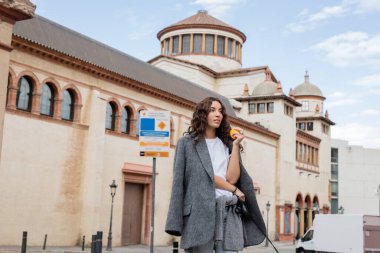İspanya 'nın Barselona şehrinde tarihi bir dönüm noktasına bakarken elinde taze turuncu ve tasma tutan gri ceketli genç kıvırcık kadın. 