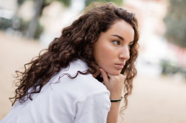 Portrait of young curly and pensive woman in white t-shirt looking away and holding hand near chin while standing on blurred urban street in Barcelona, Spain  clipart