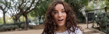 Portrait of shocked young and curly woman in white t-shirt looking at camera with open mouth while spending time in blurred park at daytime in Barcelona, Spain, banner  clipart